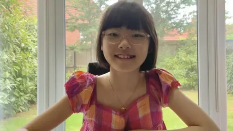 La niña es una niña con una longitud de cabello oscuro, gafas y rosa, y mantiene la cadena de oro, y se ríe del maestro. El jardín detrás de la ventana puede estar detrás de él.