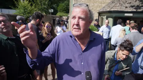 Clarkson fotos en la multitud y micrófono antes que él del periodista que no se encuentra. Lleva un abrigo morado, cabello blanco y señalando algo desde la distancia.