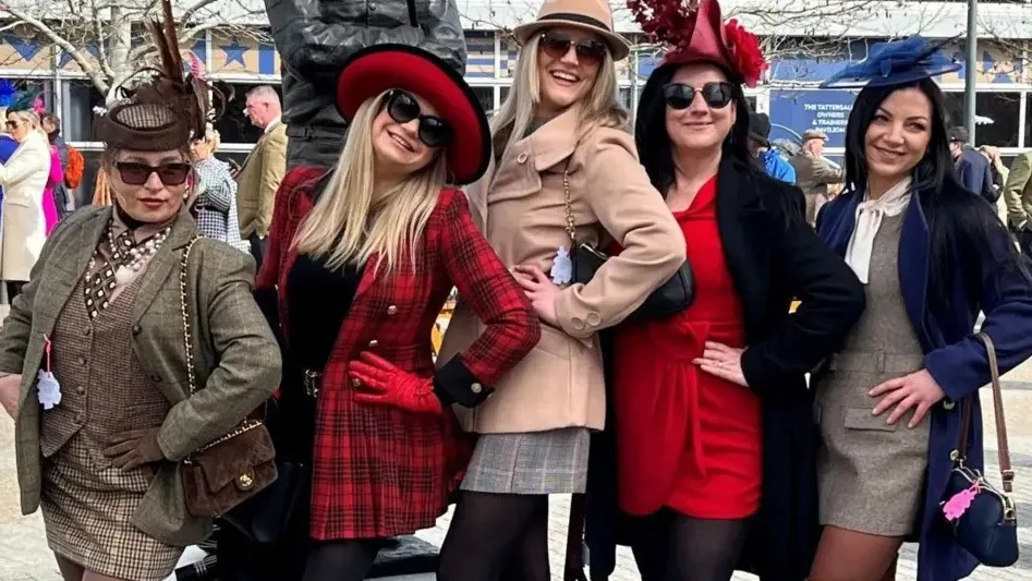 Cinco mujeres se encuentran en el hipódromo de Cheltenham. Todos están vestidos con pequeños vestidos, camisas, chaquetas y abrigos, y llevan sombreros, dijes y gafas de sol.