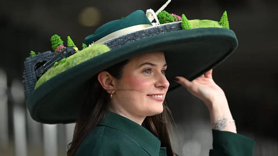 Una mujer de largo cabello negro y una camisa verde oscuro que lleva un sombrero de gran tamaño decorado con un ala y una vía de tren. Una máquina sale del túnel del capó.