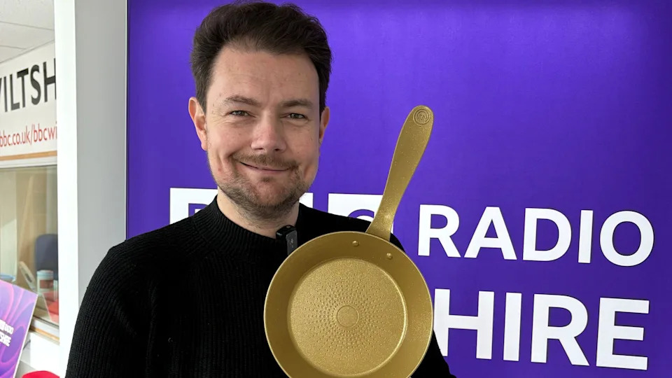 Thomas Frake, un hombre de cabello negro y barba incipiente, vestía de negro y sostenía una sartén dorada. Hay un cartel detrás de él que dice "BBC Radio Wiltshire".