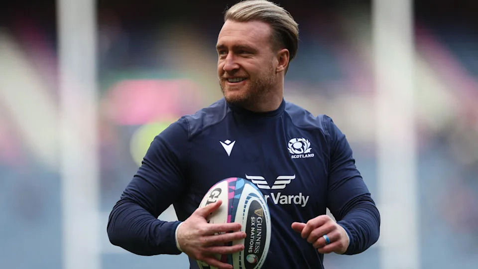 Stuart Hogg con el uniforme azul de Escocia sostiene una pelota de fútbol. Tenía el pelo rubio peinado hacia atrás.