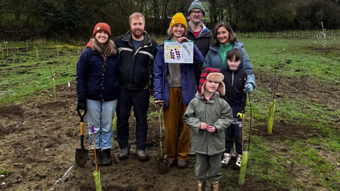 Siete personas, entre ellas dos niños, sonríen a la cámara en un campo donde se han plantado muchos árboles jóvenes. Todos visten ropa de invierno para exteriores, algunos llevan gorros con pompones y dos de ellos sostienen palas. 
